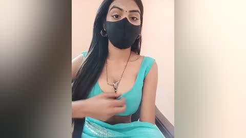 Media: Video of a South Asian woman with long black hair, wearing a black mask, turquoise bra, and a metallic skirt, adjusting her bra, indoors against a beige wall.