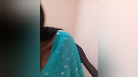 Media: Video of a woman with a light brown complexion, wearing a turquoise saree with white polka dots, partially obscured by a black hair clip, standing in a dimly lit room with neutral-colored walls.