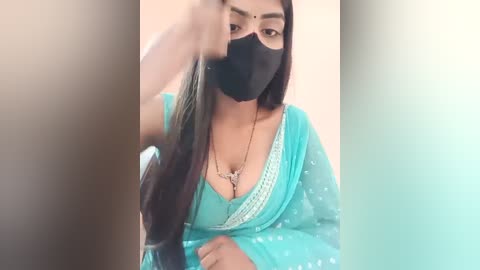 Media: Video of a young South Asian woman in a turquoise saree, black mask, and long black hair. She's seated, holding her hair up, with a pendant necklace visible. The background is blurred.