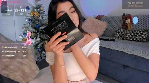 Media: A young Asian woman with long black hair, wearing a white off-shoulder top, takes a selfie video with a black compact mirror. The background shows a Christmas tree and a gray couch with a plush pillow.