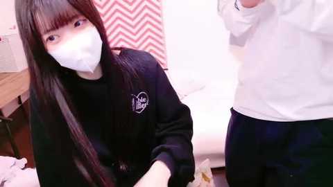 Media: Video of a young Asian woman with long black hair, wearing a black sweater, white mask, and a white surgical gown, kneeling beside a man in a white shirt and dark pants in a dimly lit room with a red and white zigzag-patterned wall.