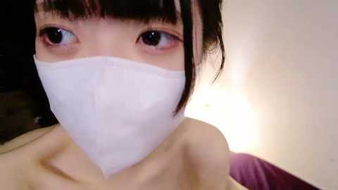 Media: A close-up video of an Asian woman with fair skin, black hair, and bangs. She wears a white surgical mask covering her nose and mouth, and her large, expressive brown eyes are visible. The background is a soft, neutral-toned wall with a warm, ambient light source creating a gentle glow.