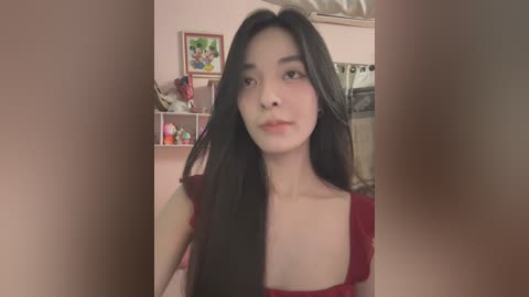 Media: Video of a young Asian woman with long black hair, wearing a red top, standing indoors in a kitchen with shelves, framed art, and cooking utensils visible in the background.