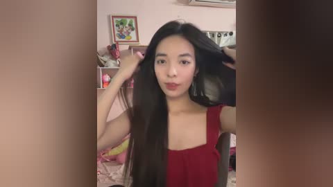 Media: Video of a young Asian woman with long black hair, wearing a red sleeveless top, sitting in a room with pink walls, shelves filled with books and toys, and a framed painting.