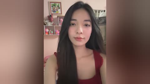 Media: Video of a young Asian woman with long black hair and fair skin, wearing a red sleeveless top, standing in a cozy, pink-themed room with shelves filled with plush toys.