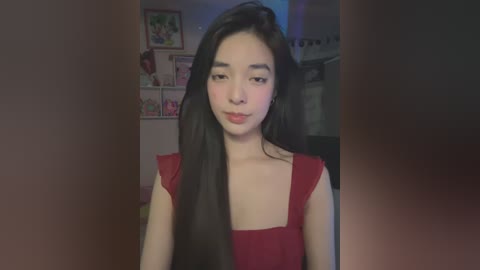 Media: Video of an East Asian woman with long black hair, fair skin, and wearing a red dress, standing in a dimly lit, cluttered room with framed pictures and books on shelves.