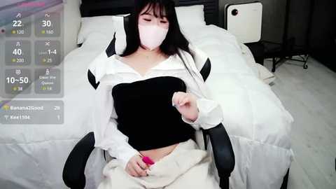 Media: A video of a woman with long black hair, wearing a white mask, black top, and beige pants, sitting in a white chair, holding a pink vibrator, in a bedroom with a white bed and grey furniture.