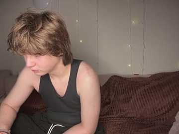 Media: Video of a young boy with light brown hair, wearing a black tank top, sitting on a bed with a brown quilt, looking down thoughtfully. Background features a string of fairy lights and a plain white wall.