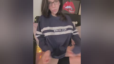 A video of a young woman with glasses and dark hair, wearing a navy and white striped sweater with \"WHITE FOX\" on it, lifting it to reveal her bare thighs. Background features a red lipstick-shaped object.