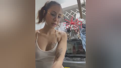 Media: Video of a young Asian woman with glasses, wearing a white tank top, sitting near a glass vase with red flowers. Background shows a partially visible kitchen with a sink.