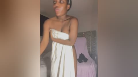 Media: Video of a slender, dark-skinned woman with short hair wrapped in a white towel, standing in a dimly lit bedroom with a bed, a black garment on the floor, and beige walls.