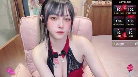 Media: Video of an Asian woman with pale skin, black hair, wearing a black lace bralette and red necklace, sitting on a pink cushioned chair. Background shows a wooden cabinet.