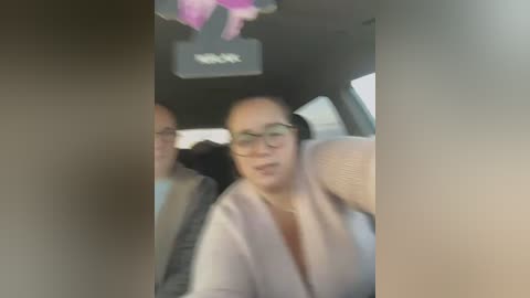 Media: A blurred video of an Asian woman with glasses and a beige cardigan sitting in a car, partially obstructed by the vehicle's interior.