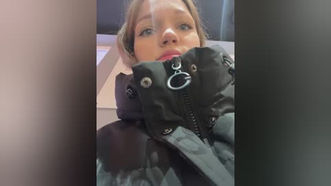 Media: Video of a young Caucasian woman with fair skin, light brown hair, and light blue eyes, wearing a black quilted jacket with a metal zipper and circular logo, sitting in a dimly lit room.