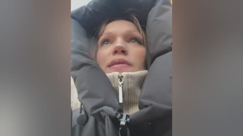 Media: Video of a woman in a puffy black winter coat, looking upwards, with her face partially obscured by the coat's high collar, set against a blurred, neutral background.