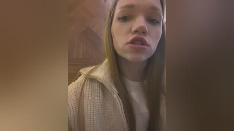 Media: A video of a young woman with light skin and long brown hair, wearing a beige knit sweater over a white shirt, standing in a dimly lit, rustic room with wooden walls and beams.