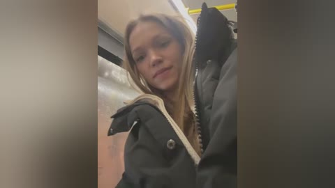 Media: A video of a young Caucasian woman with long blonde hair and light skin, wearing a black puffer jacket, standing in a subway car. The background shows metallic subway railings and a blurred interior.