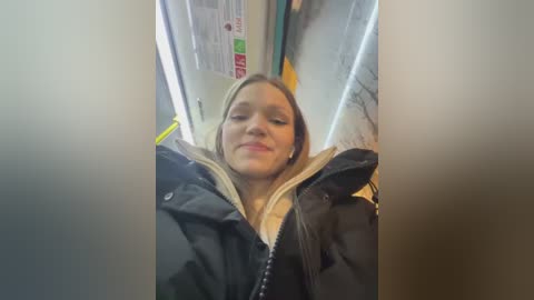 Video of a young woman with light skin and long blonde hair, smiling inside a subway train, wearing a black jacket with a hood.