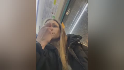 Media: Video of a young woman with long blonde hair, wearing a black jacket, sitting on a subway train. Her mouth is partially covered by her hand. The background shows other passengers, advertisements, and a tiled floor.