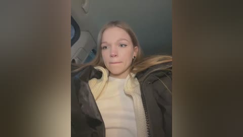 Media: A video of a young Caucasian woman with light brown hair, wearing a black jacket over a white sweater, sitting in a car. The background shows part of the vehicle's interior.