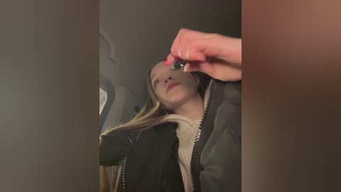 Media: Video of a young woman in a car, wearing a black jacket and a beige scarf, holding a phone in her right hand, looking up.