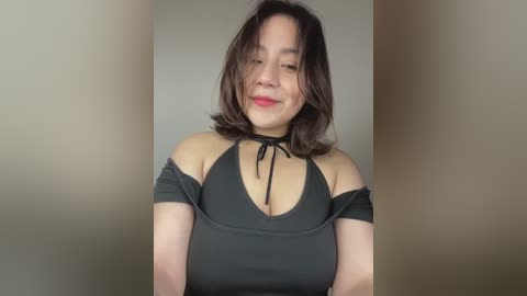 Media: Video of an East Asian woman with shoulder-length dark hair, wearing a black halter top that accentuates her medium-sized breasts. She smiles softly against a plain beige background.