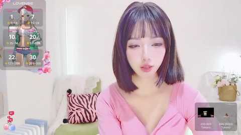 Media: Video of a young East Asian woman with straight, shoulder-length black hair, fair skin, and light makeup, wearing a pink cardigan, sitting in a bright, minimalist living room with floral accents.