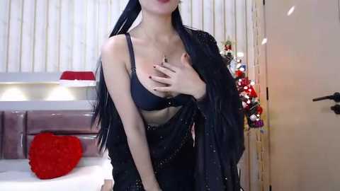 Media: Video of an Asian woman with long black hair, wearing a black bra, and a sheer black robe, standing in a modern bedroom with white vertical blinds, red heart pillow, and Christmas decorations.