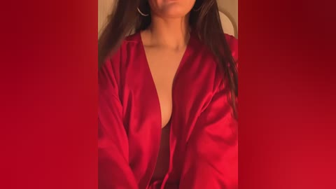 Media: Video of a woman with long brown hair, wearing a red satin robe with a deep V-neck, revealing cleavage. She has large hoop earrings and stands against a solid red background.