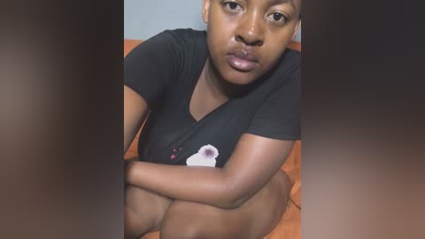 Media: Video of a young Black woman with medium skin tone, sitting on a wooden floor, wearing a black t-shirt, with a white flower graphic, and a neutral expression.