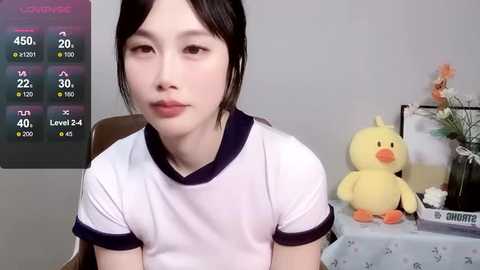 Media: Video of a young Asian woman with pale skin, black hair, and a slender build, wearing a white t-shirt. Background features a duck toy and a floral arrangement on a table.