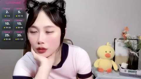 Media: Video of an Asian woman with fair skin, wearing black cat ears and a white shirt, resting her head on her hand, surrounded by a digital clock, a stuffed duck, and a small bouquet.