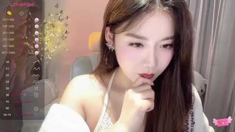 Media: A young East Asian woman with long brown hair, fair skin, and soft makeup, wearing a white off-shoulder top, poses thoughtfully. The background features a pink wall with a lit lamp and a plush toy.