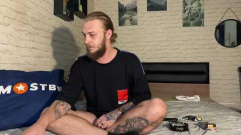 Media: Video of a bearded man with long hair, tattoos, wearing a black long-sleeve shirt, sitting cross-legged on a bed in a dimly lit room with white brick walls.