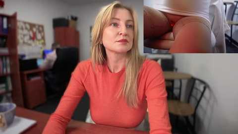 Video of a blonde woman in a red sweater, seated at a desk, with a close-up of a hand holding a red object. Background includes bookshelves and a computer monitor.