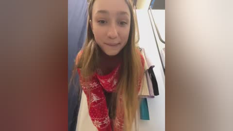 Media: Video of a young, light-skinned girl with long, straight, blonde hair, wearing a red and white patterned scarf, leaning forward with a gentle smile, in a bathroom with a blue towel and hanging clothes in the background.