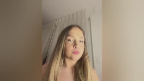 Media: A blurry video of a young Caucasian woman with long, blonde hair and fair skin, standing in a dimly lit room with beige walls and a white curtain.