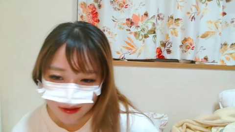 Media: Video of an Asian woman with straight, shoulder-length brown hair wearing a white surgical mask, red lipstick, and a white top, sitting indoors with floral curtains and a beige wall in the background.