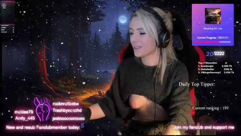 Media: Video of a live stream featuring a blonde woman in headphones, sitting in a dark, cozy room with a fireplace and a forest background.