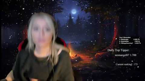 Media: Video of a woman with long, blonde hair, wearing a black hoodie, sitting in front of a campfire in a forest at night, surrounded by glowing red logs and a full moon.