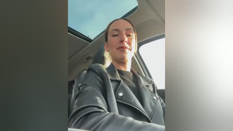 Media: Video of a fair-skinned woman with straight blonde hair, wearing a black leather jacket and black top, seated in a car with a clear blue sky visible through the sunroof.