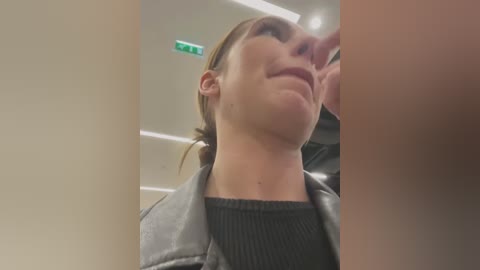 Media: Video of a woman with fair skin, brown hair, and a slight smile, wearing a black top and gray jacket, taken from a low angle in an indoor setting with a green exit sign and fluorescent lights.