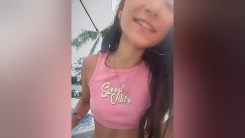 Media: A video of a young woman with long dark hair, fair skin, and a slim physique, wearing a pink crop top with \"Good Vibes Only\" text, smiling outdoors.