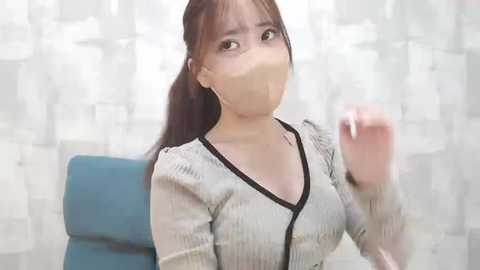 Media: Video of an Asian woman with long brown hair, wearing a beige surgical mask, blue shirt, and black sweater, seated indoors with white curtains in the background.