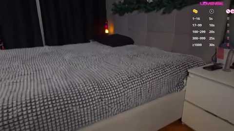 Video of a modern bedroom with a white bed, black and white checkered duvet, dark curtains, and a small white nightstand with a lamp and bottle.