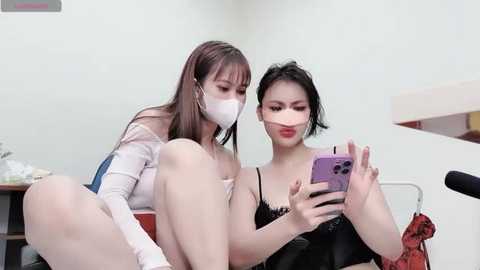 Media: Video of two East Asian women taking a selfie. One wears a white mask, long sleeves, and a skirt, while the other has short black hair, red lipstick, and a black top. Background shows a studio setting with a camera.