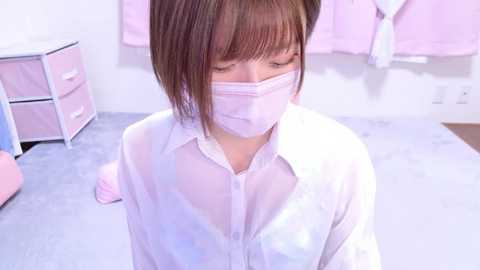 Media: Video of an Asian woman with short brown hair, wearing a white button-up shirt and a pink face mask, kneeling on a light gray carpeted floor. Background includes pink storage bins and a pink towel hanging on a wall hook.