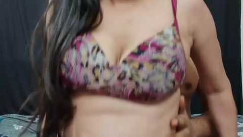 Media: Video of a woman with long black hair wearing a floral-patterned pink bra, revealing a fit, slightly muscular physique, with a blurred background.