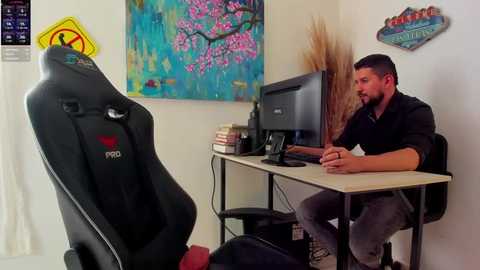 Media: Video of a bearded man in a black shirt and gray pants, sitting at a desk with a computer monitor, surrounded by colorful wall art, including a cherry blossom painting, in a modern office space.