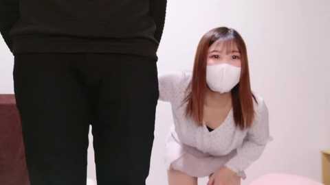 Media: Video of an Asian woman in a white mask, kneeling with a submissive expression, facing a person wearing black pants, in a minimalistic room.